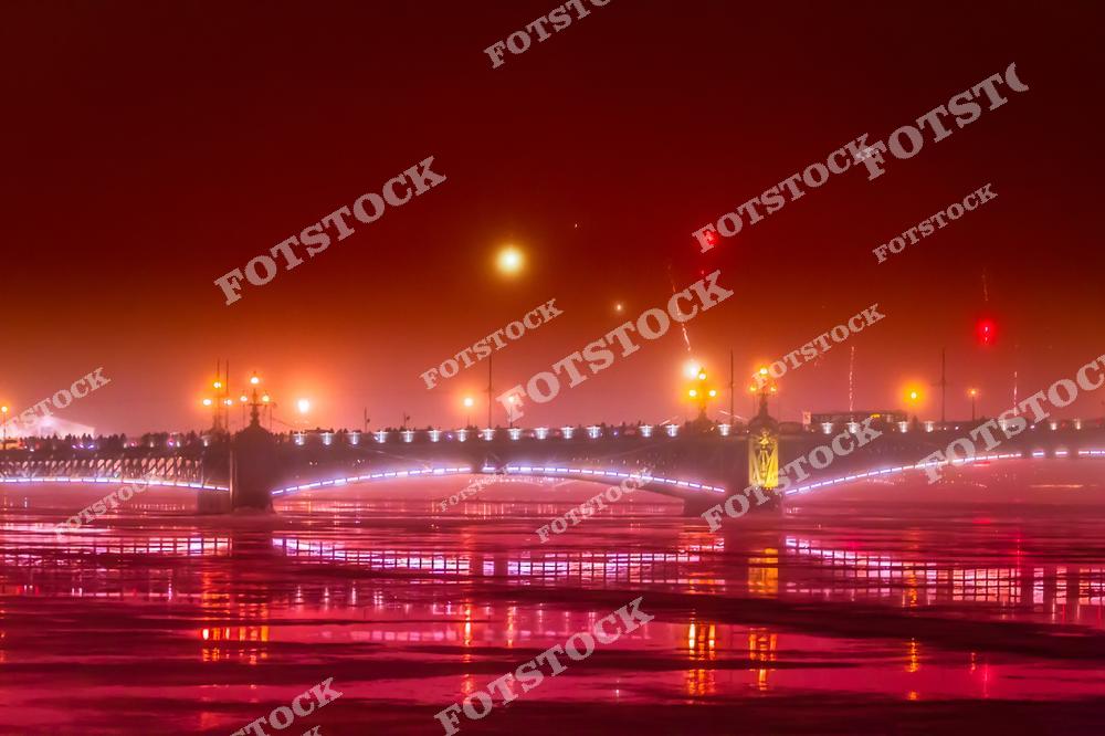 На фотографии изображён Троицкий мост, один из самых известных мостов Санкт-Петербурга. Яркая подсветка подчёркивает изящество архитектуры моста, а её отражение играет на льду и воде Невы. Туман и ночное освещение создают романтическую и загадочную атмосферу. Этот мост соединяет центр города с Петроградской стороной и является важной частью городского ландшафта.